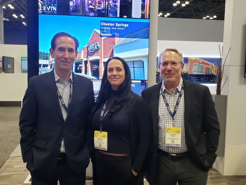 Two men and a woman standing at Levin Management's booth with a large photo of a shopping center in the background