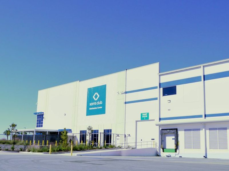 Large, two-story Sam's Club warehouse under a blue Florida sky with palm trees in the distant background
