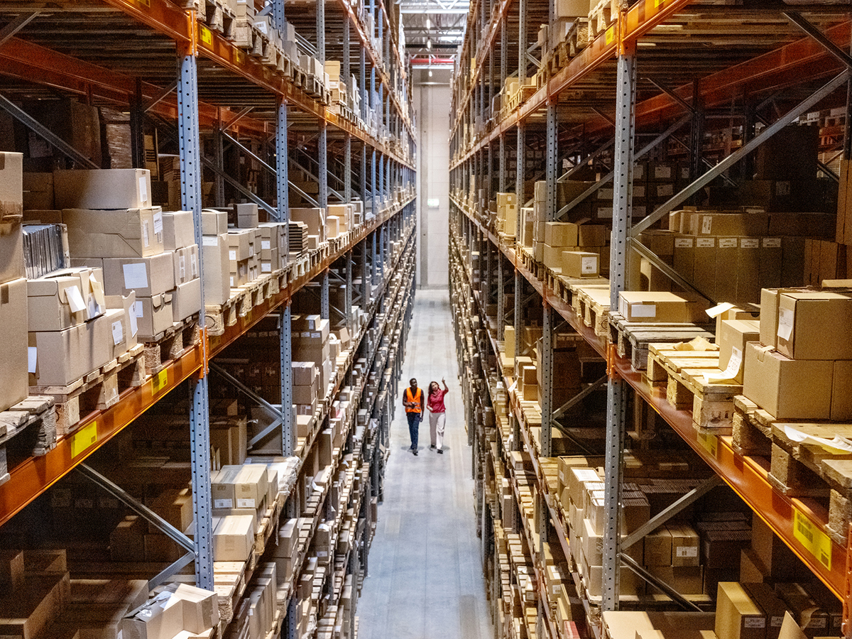 High angle view of a warehouse manager walking with foremen checking stock on racks. Businesswoman discussing stock with a male worker while walking by racks in warehouse.