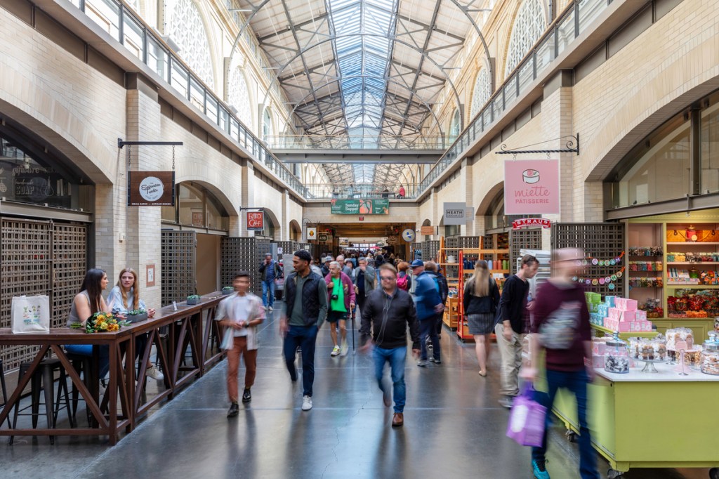 People walking inside the central nave of the Ferry Building, with skylight and small stores on both sides