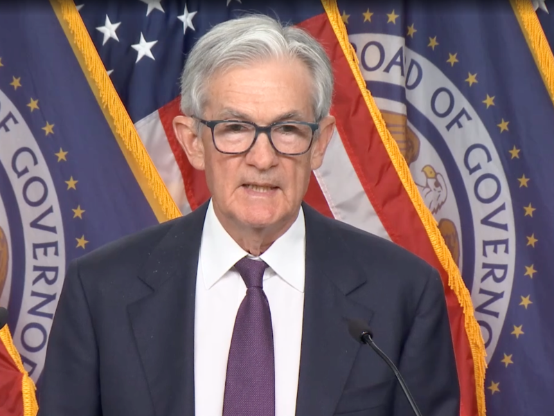 Silver-haired man in blue suit and tie stands in front of flags