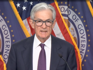 Silver-haired man in blue suit and tie stands in front of flags