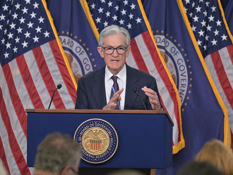 Silver-haired man in blue suit and tie standing on podium behind American flags and Federal Reserve flags