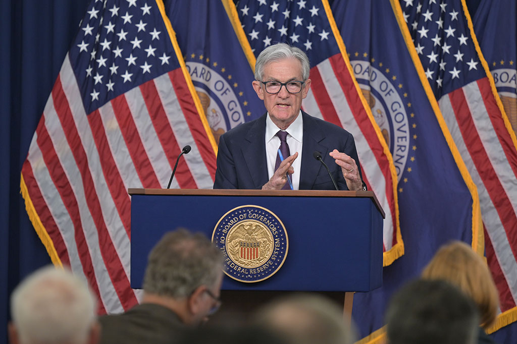 Silver-haired man in blue suit and tie standing on podium behind American flags and Federal Reserve flags