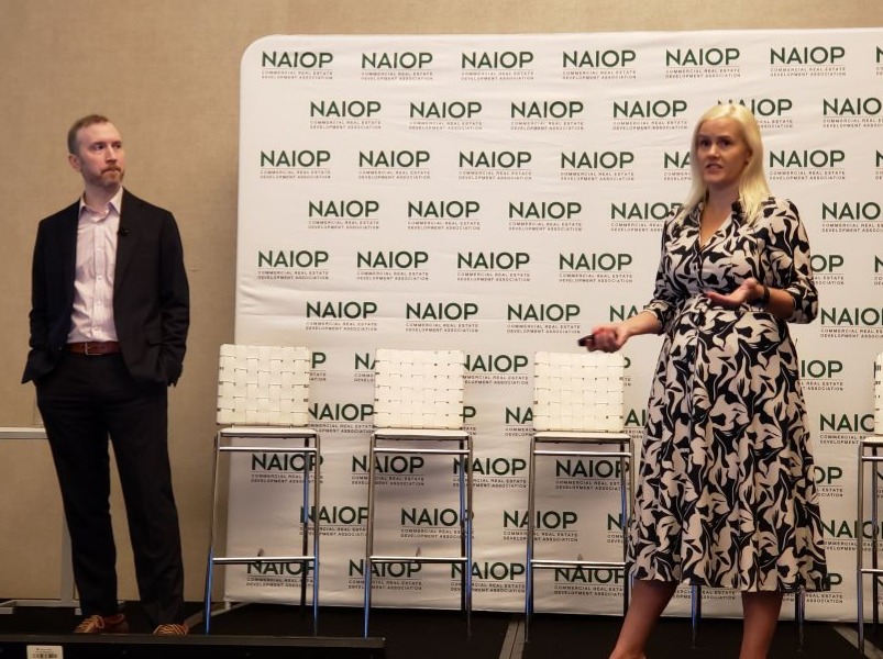 woman speaking in front white chairs and large white sign with NAIOP; man stands listening at left with hands in pockets