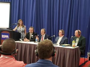 At left, woman stands behind lectern with BOMA sign. To her left, four men wearing headsets are seated at a table