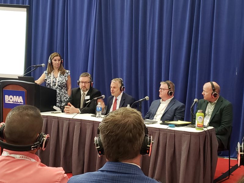 At left, woman stands behind lectern with BOMA sign. To her left, four men wearing headsets are seated at a table 
