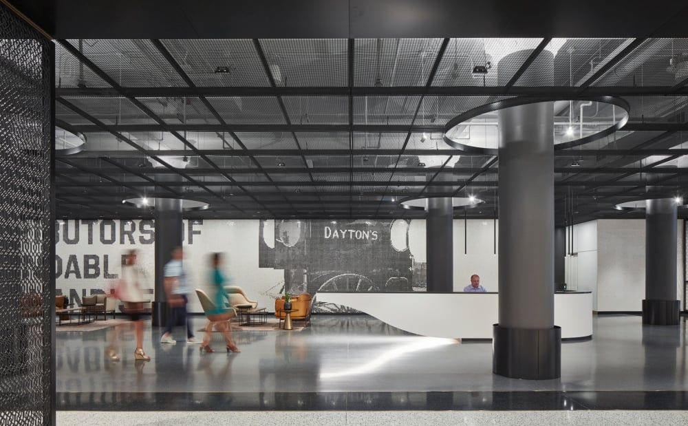 Modern lobby with black grid ceiling and large columns. Blurred motion of people walking, a person seated at a white reception desk. Calm ambiance.