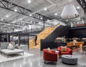 Modern open office space with high ceilings, featuring a wooden staircase, gray and red seating, and people engaged in conversation. Contemporary and inviting atmosphere.