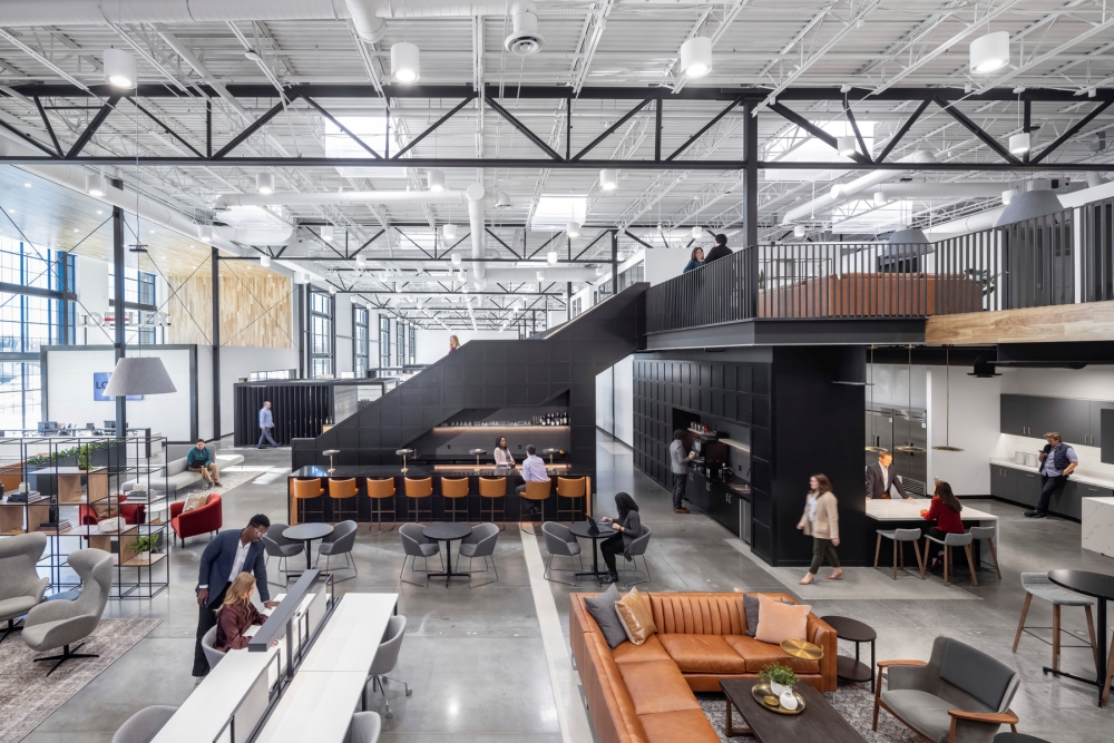 Spacious modern office with people collaborating. Features open seating, a central staircase, and lofted area. Bright, airy space with sleek design.