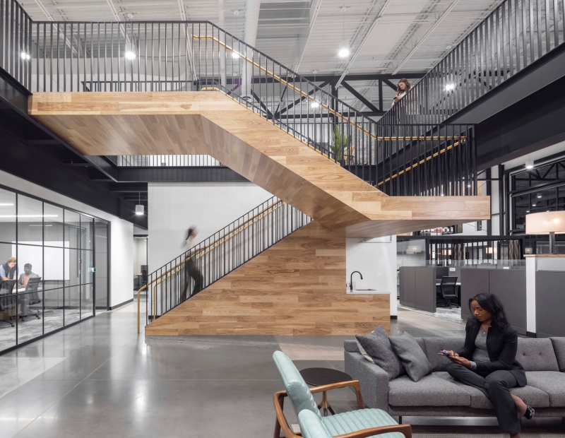 Modern office interior with a wooden staircase at the center. A woman sits on a gray sofa using a phone. Glass-walled meeting room visible on the left.