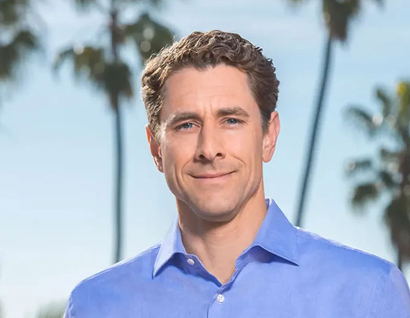 Smiling, brown-haired man wearing open-necked blue shirt with palm trees in the background.