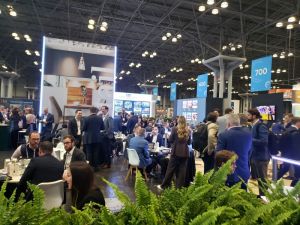 People in suits sitting at round tables in front of large displays and banners under a high convention center ceiling
