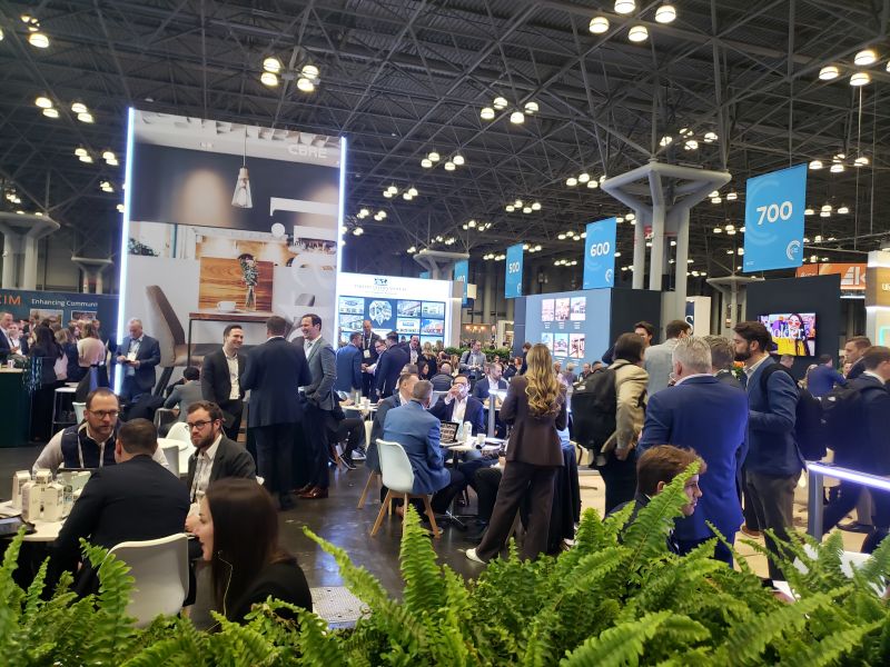 People in suits sitting at round tables in front of large displays and banners under a high convention center ceiling