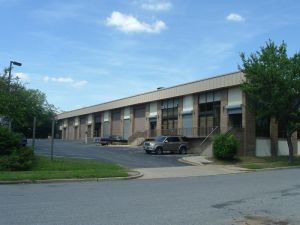 Single-story warehouse with neutral beige trim. In foreground, small driveway flanked by two trees