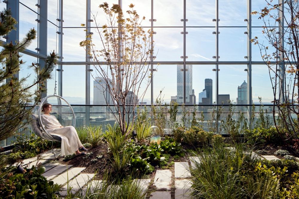A woman is seated in a chair near a large window, with an array of plants adding a touch of nature to the indoor space.