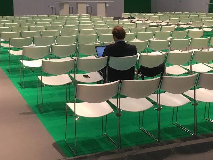 Man sits with laptop in lap, alone in a conference hall full of white chairs