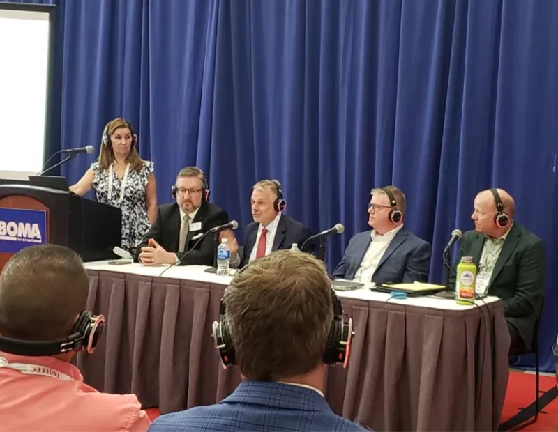 At left, woman stands behind lectern with BOMA sign. To her left, four men wearing headsets are seated at a table