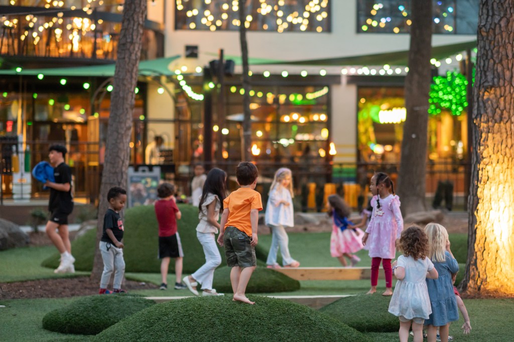 Exterior shot of children playing around a green space, with lights hanging around trees