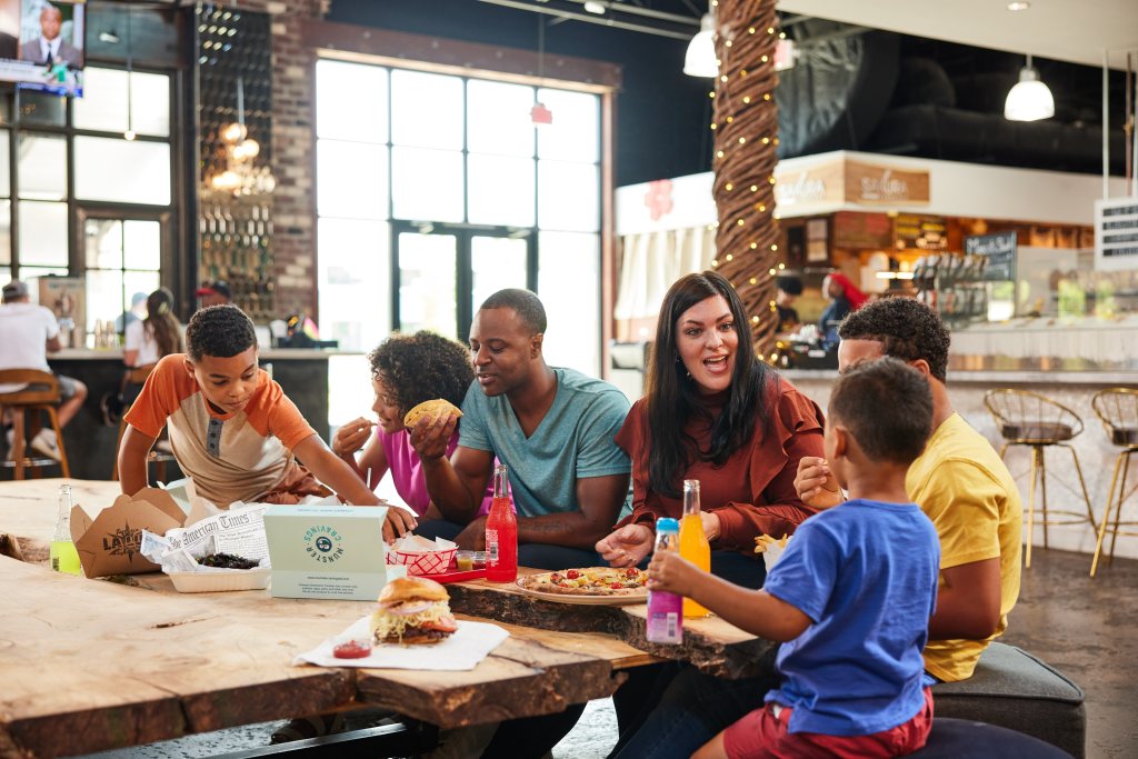 Interior shot of children and adults sitting around at a table, while socializing and eating.