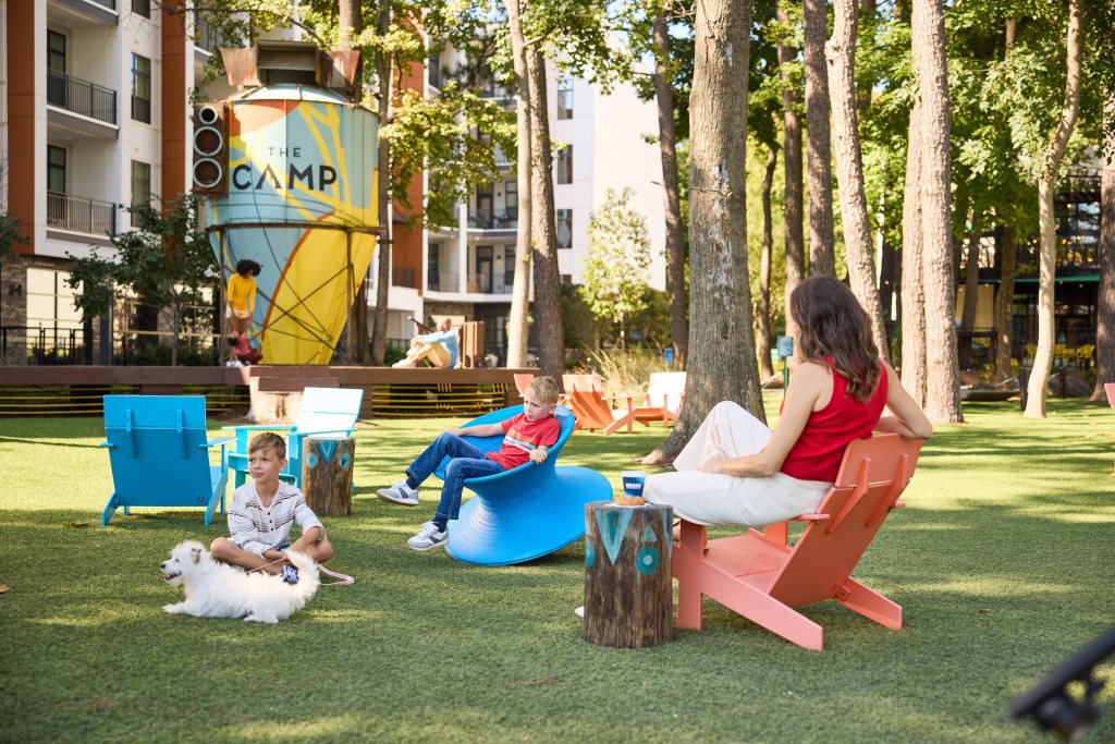 Children are playing in an outdoor area, with adults in the space as well, either standing up or sitting down in chairs. A dog is next to a child.
