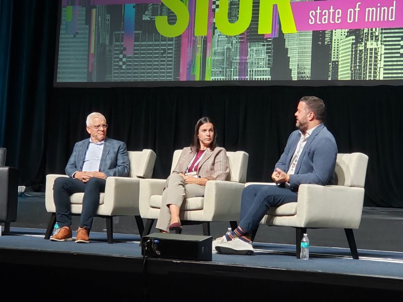 Woman in beige suit flanked by two men wearing light blue blazers, seated on white chairs on a stage and in front of a sign saying SIOR state of mind