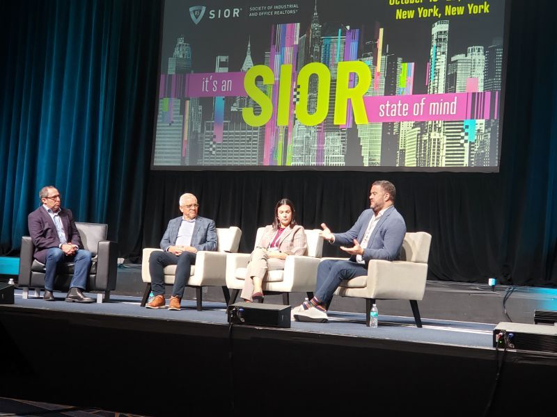 Three men and one woman seated in white chairs on a stage in front of a sign saying, It's an SIOR state of mind.