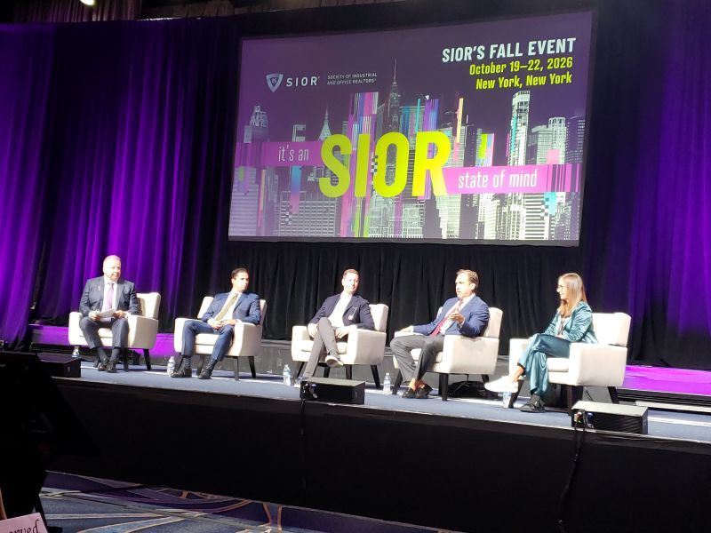 Four men and one woman seated in white chairs on a stage in front of a purple curtain and a sign saying "It's an SIOR state of mind."
