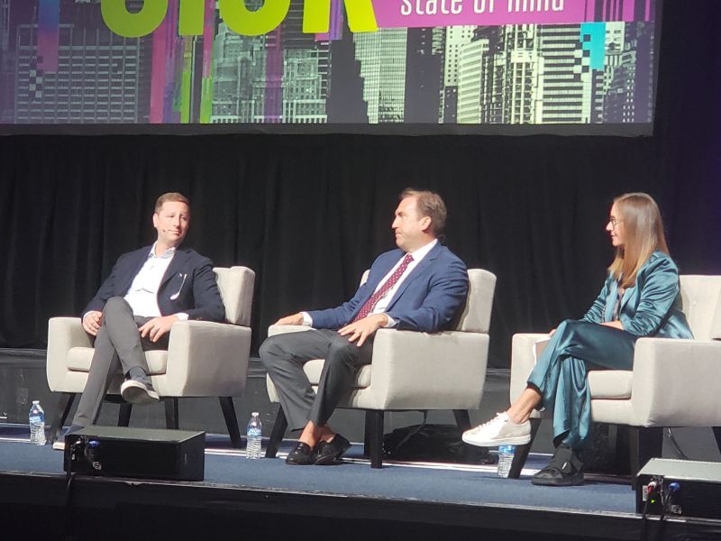 Two men and one woman wearing business attire seated in white chairs on a stage.