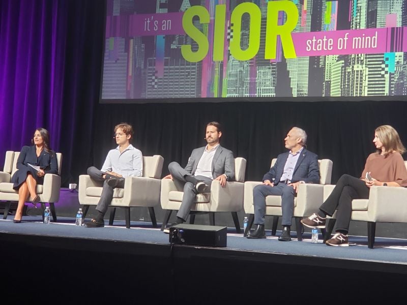 Four people in business attire sitting in white chairs on a stage in front of a sign that says, It's an SIOR state of mind