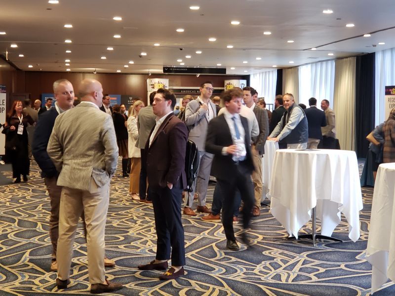 People in business attire talking in a hotel reception area with small round tables covered by white tablecloths on the right.