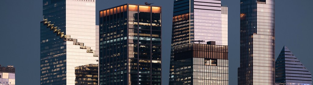 A dense cluster of Manhattan office towers fills the frame, showcasing the reflective interplay of glass, steel, and fading daylight. This detailed look at NYC architecture highlights texture, repetition, and scale.